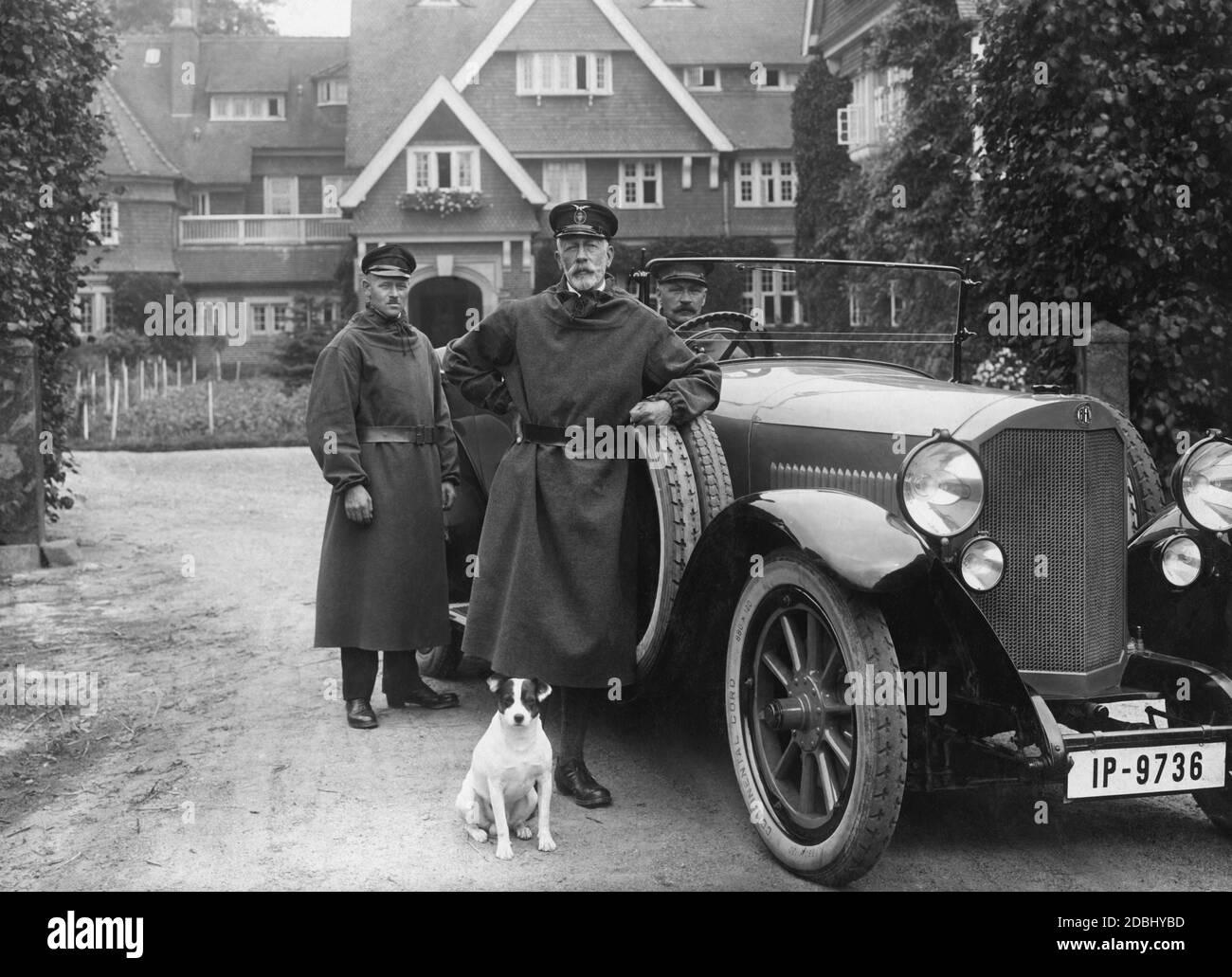 Prinz Heinrich von Preußen (Mitte) lehnt sich an seinen Benz 11. Er trägt eine Mütze mit dem Logo des Kaiserlichen Automobil-Clubs. Sein Hund sitzt vor ihm. Das Auto hat ein preußisches Kennzeichen aus dem Bundesland Schleswig-Holstein, das mit IP beginnt. Im Hintergrund befindet sich das Herrenhaus Hemmelmark, in dem Heinrich von Preußen lebte. Stockfoto