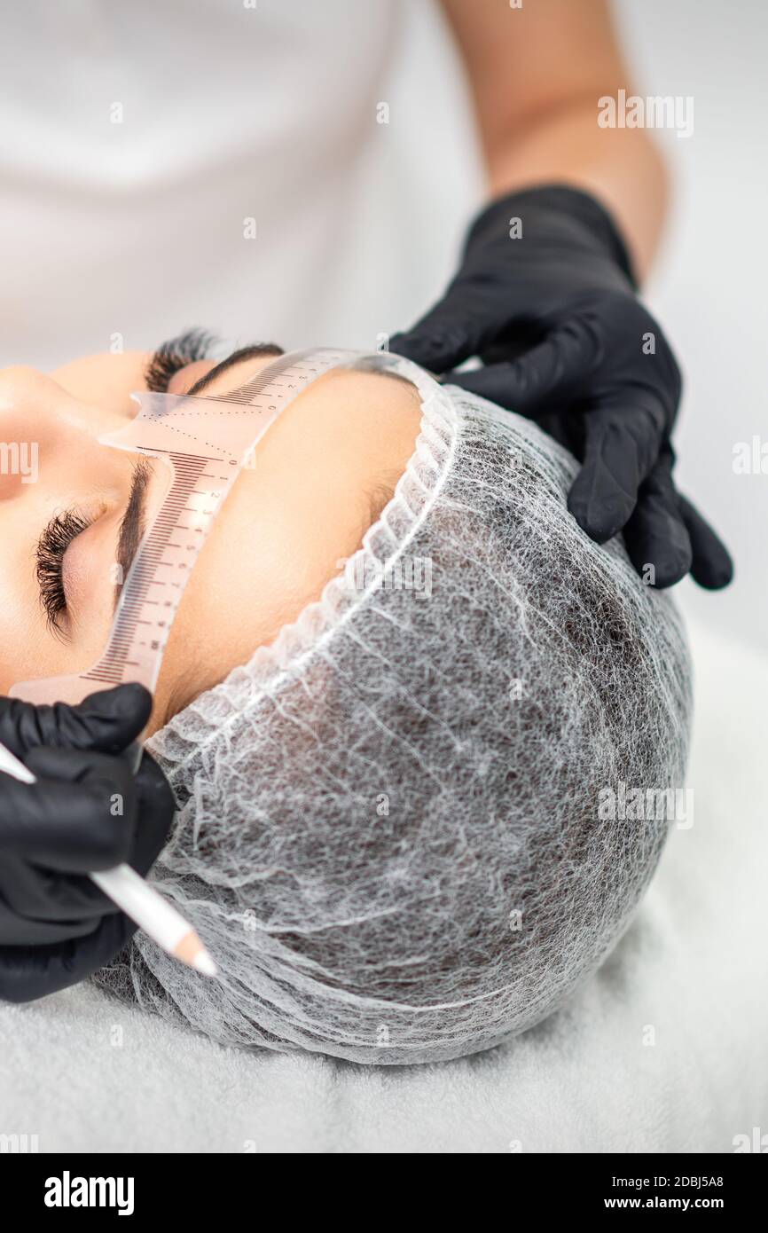 Der Kosmetologe misst mit Lineal die Brauen des jungen Kaukasiers Frau vor Permanent Make-up Tattoo Stockfoto