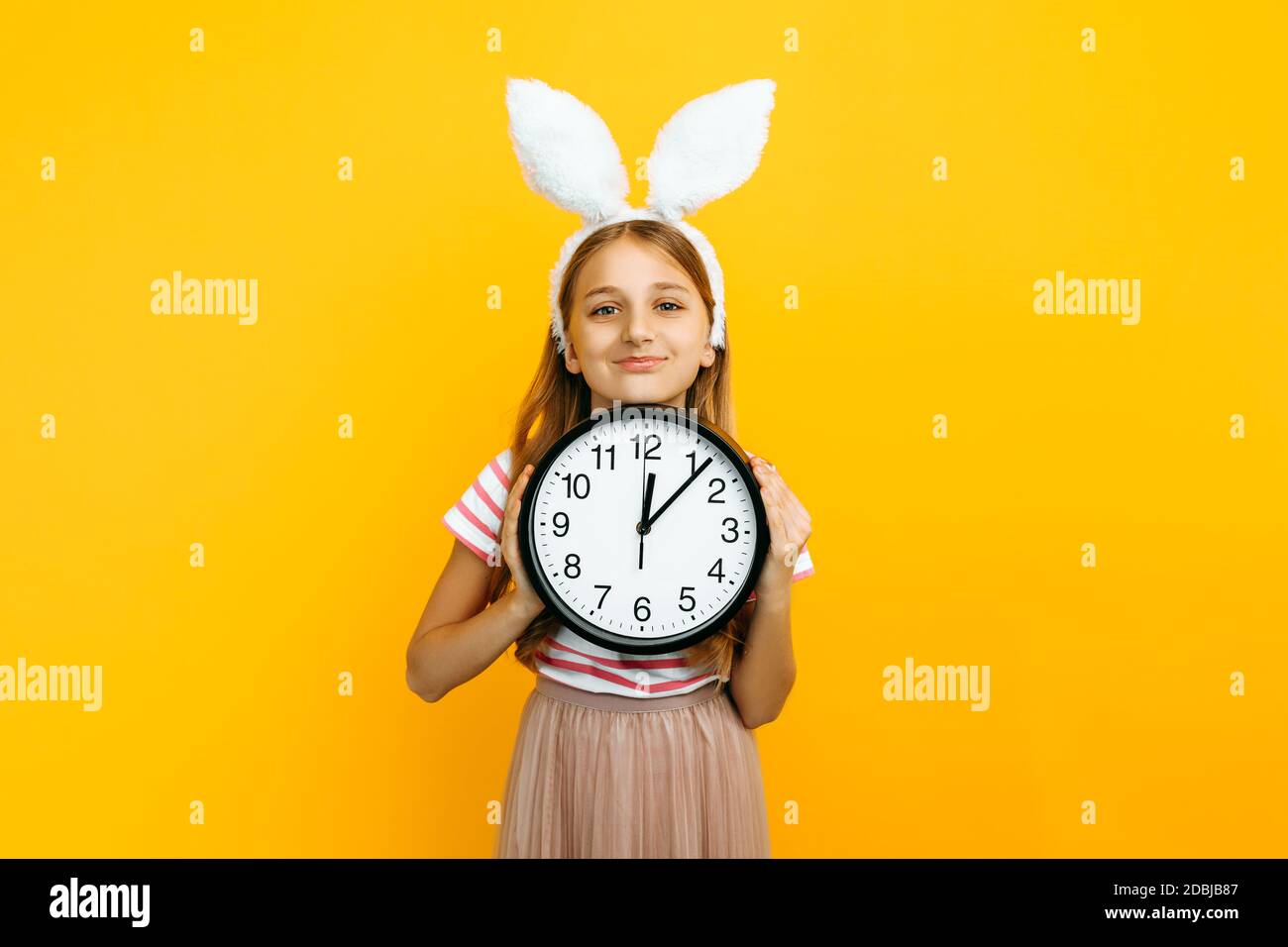 Happy schönes Mädchen auf dem Kopf mit Kaninchenohren, mit einer Wanduhr in den Händen auf einem gelben Hintergrund. Symbol von Ostern und Frühling. Stockfoto