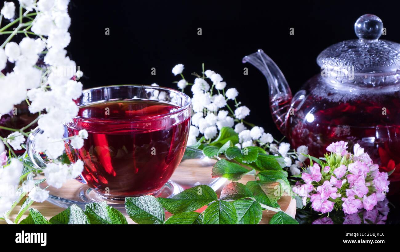 Ein Becher roten Tees und eine Teekanne in weißen Hibiskusblüten und grünen Blättern medizinischen Tees auf einem Holzständer.Zen-Tee Zeremonie. Foto von roten Kräuter Indi Stockfoto