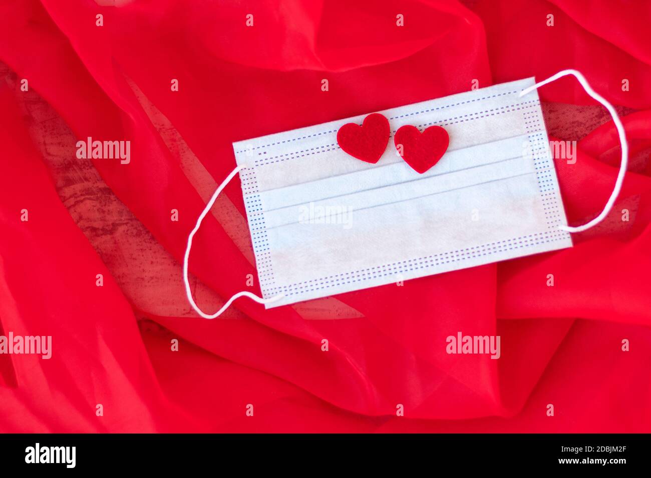Blaue medizinische Schutzmaske mit zwei roten Herzen auf rotem Hintergrund. Romantisches Geschenk zum Valentinstag. Liebe. Konzept des Heiratsvorschlags. Weichfokus. Speicherplatz kopieren. Draufsicht Stockfoto