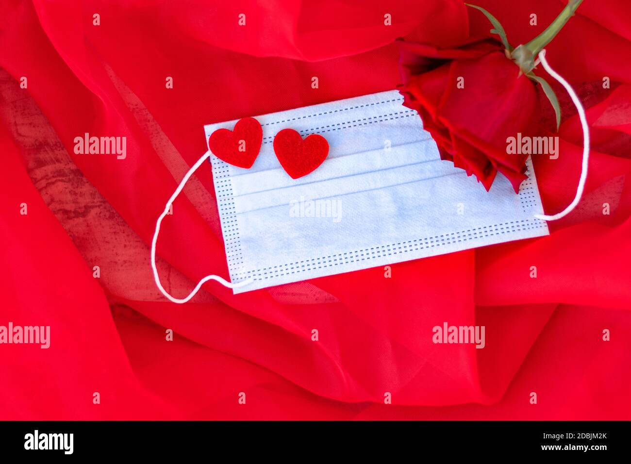Blaue medizinische Schutzmaske mit zwei roten Herzen auf rotem Hintergrund. Rote Rose neben Maske. Romantisches Geschenk zum Valentinstag. Liebe. Konzept des Heiratsvorschlags. Weichfokus. Speicherplatz kopieren. Draufsicht Stockfoto
