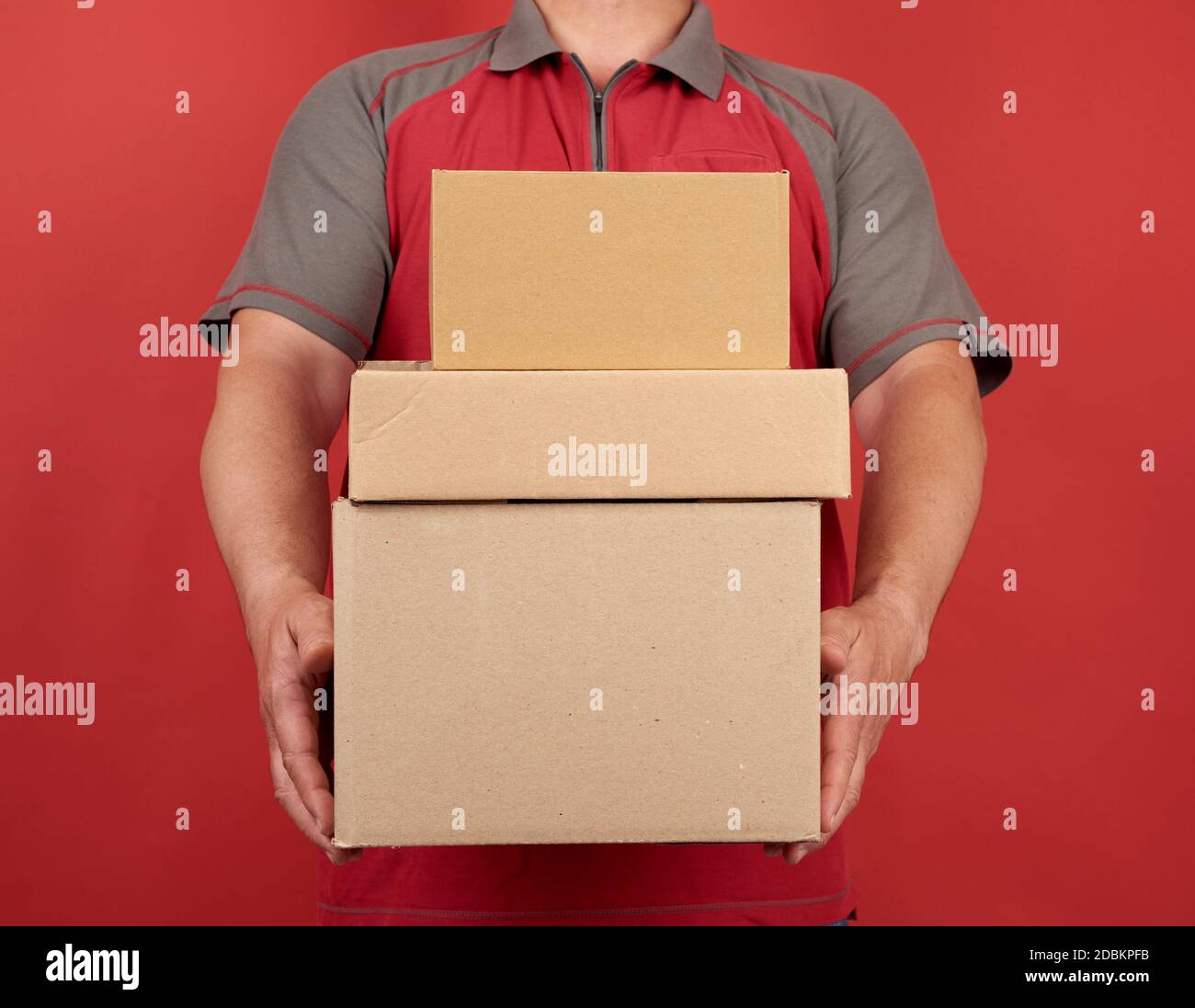 Erwachsene Mann in einem T-Shirt hält einen Stapel von großen Karton braunen Boxen auf einem roten Hintergrund, Konzept der Lieferung von Waren, bewegen Stockfoto