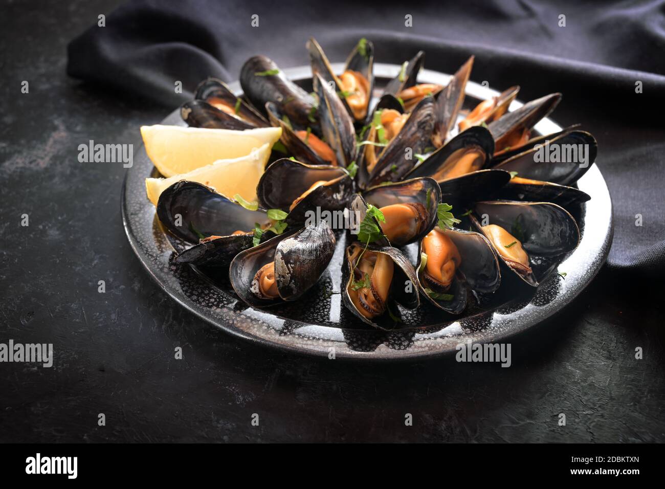 Muscheln mit Zitrone und Petersilie auf einem Teller mit dunkler Serviette auf einem schwarzen Schiefertisch, ausgewählter Fokus, sehr enge Schärfentiefe Stockfoto