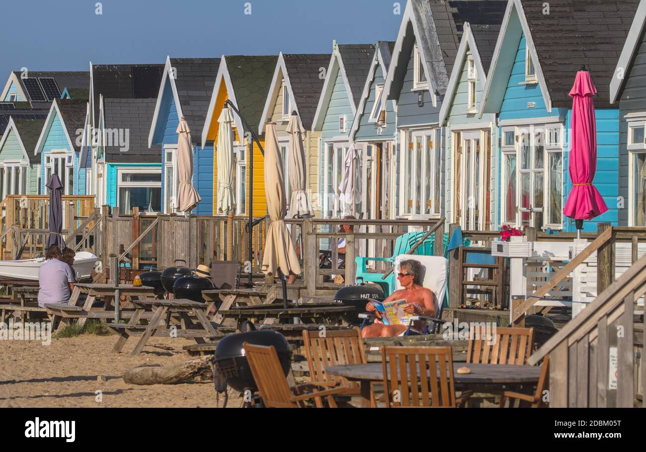 Menschen sitzen in der Sonne Sonnenbaden vor bunten Strandhütten auf Mudeford Sand Spit, Christchurch Großbritannien Stockfoto