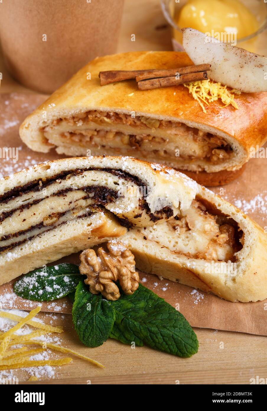 Mohn- und Apfelstrudel auf einem rustikalen Holztablett. Das Dekor der Duftrollen besteht aus Zimt, Minze, Nuss, Zitronenschale und Puderzucker. Nahaufnahme, verschwommen Stockfoto