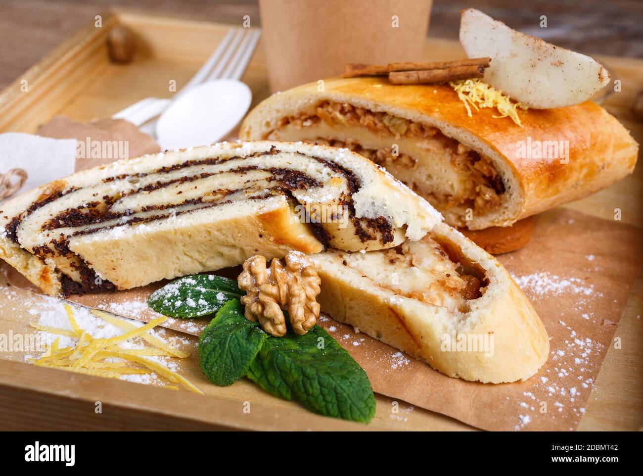 Mohn- und Apfelstrudel auf einem rustikalen Holztablett. Das Dekor der Duftrollen besteht aus Zimt, Minze, Nuss, Zitronenschale und Puderzucker. Nahaufnahme, verschwommen Stockfoto