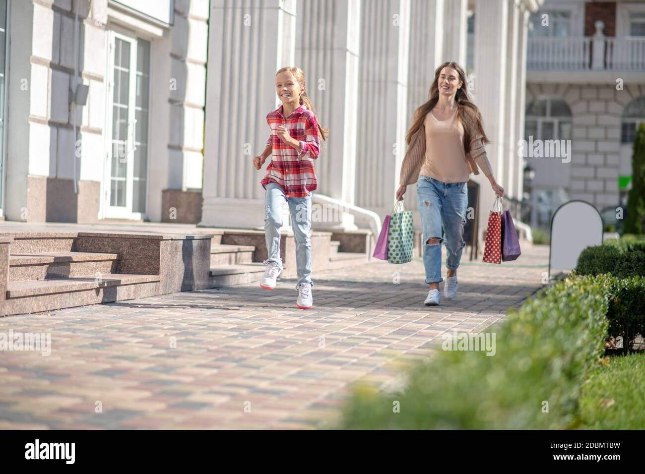 Glückliches Mädchen läuft vor ihrer Mutter zu Fuß mit Einkaufstaschen Stockfoto