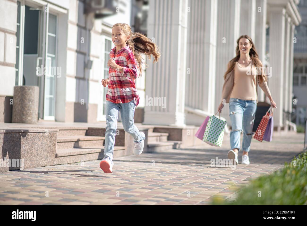 Lächelndes Mädchen läuft vor ihrer Mutter zu Fuß mit Einkaufstaschen Stockfoto