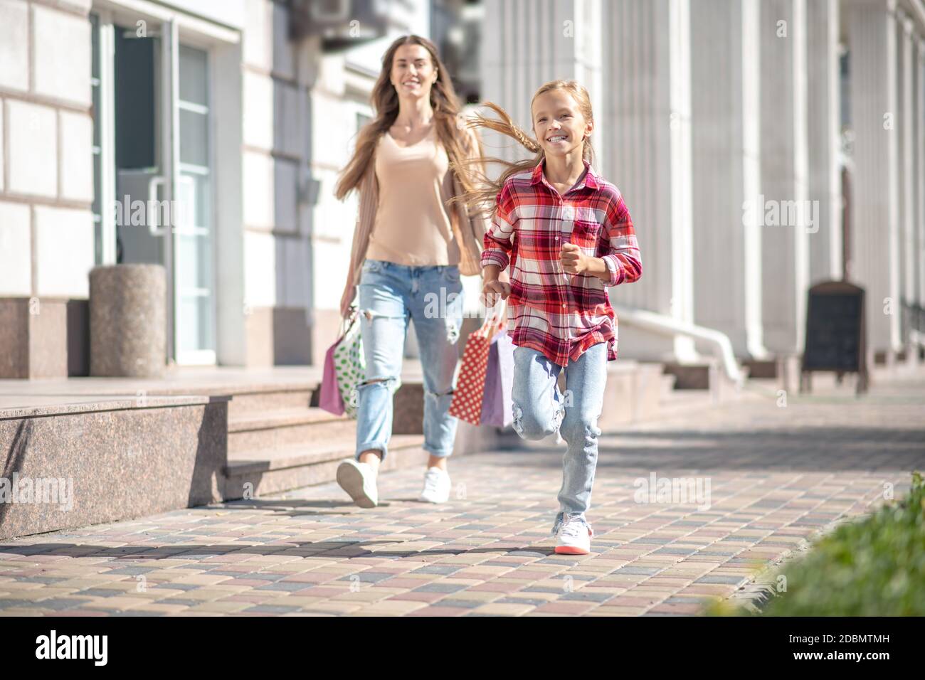 Lächelndes Mädchen mit Einkaufstasche vor ihr laufen Mama draußen Stockfoto