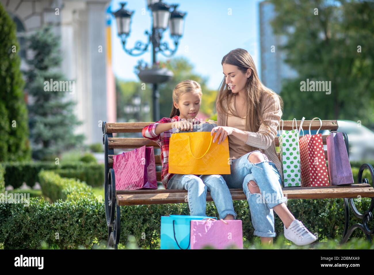 Glückliche Mutter und Tochter sitzen auf Parkbank und nehmen Artikel aus der Einkaufstasche Stockfoto