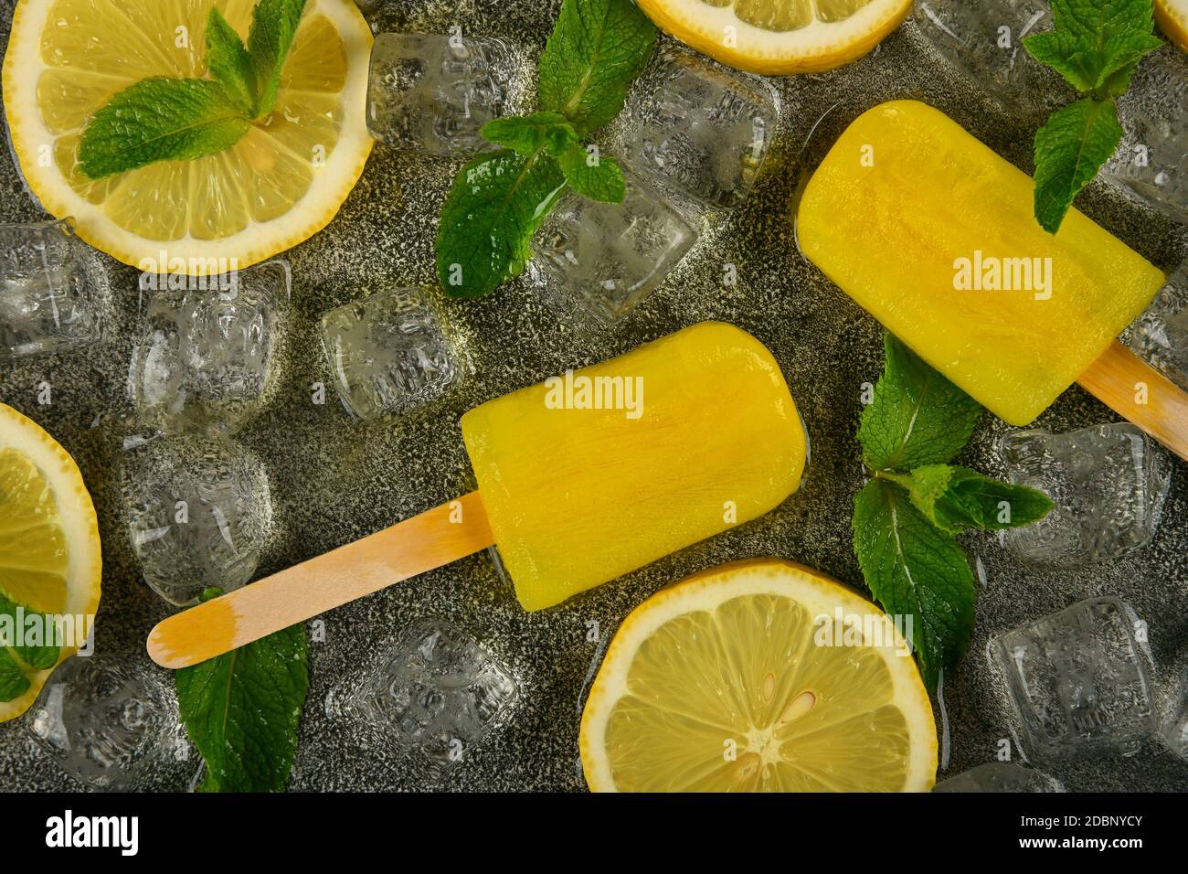 Nahaufnahme gefrorener Saft und Limoncello-Popsicles mit frischen Zitronenscheiben, grünen Minzblättern und Eiswürfeln auf grauer Tischfläche, erhöhte Ansicht von oben, dire Stockfoto