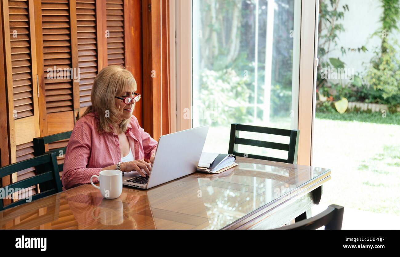 Reife Frau mit Laptop. Sie ist zuhause drinnen. Großmutter in einem Holztisch mit einem silbernen Computer. Kaffeetasse beiseite. Stockfoto
