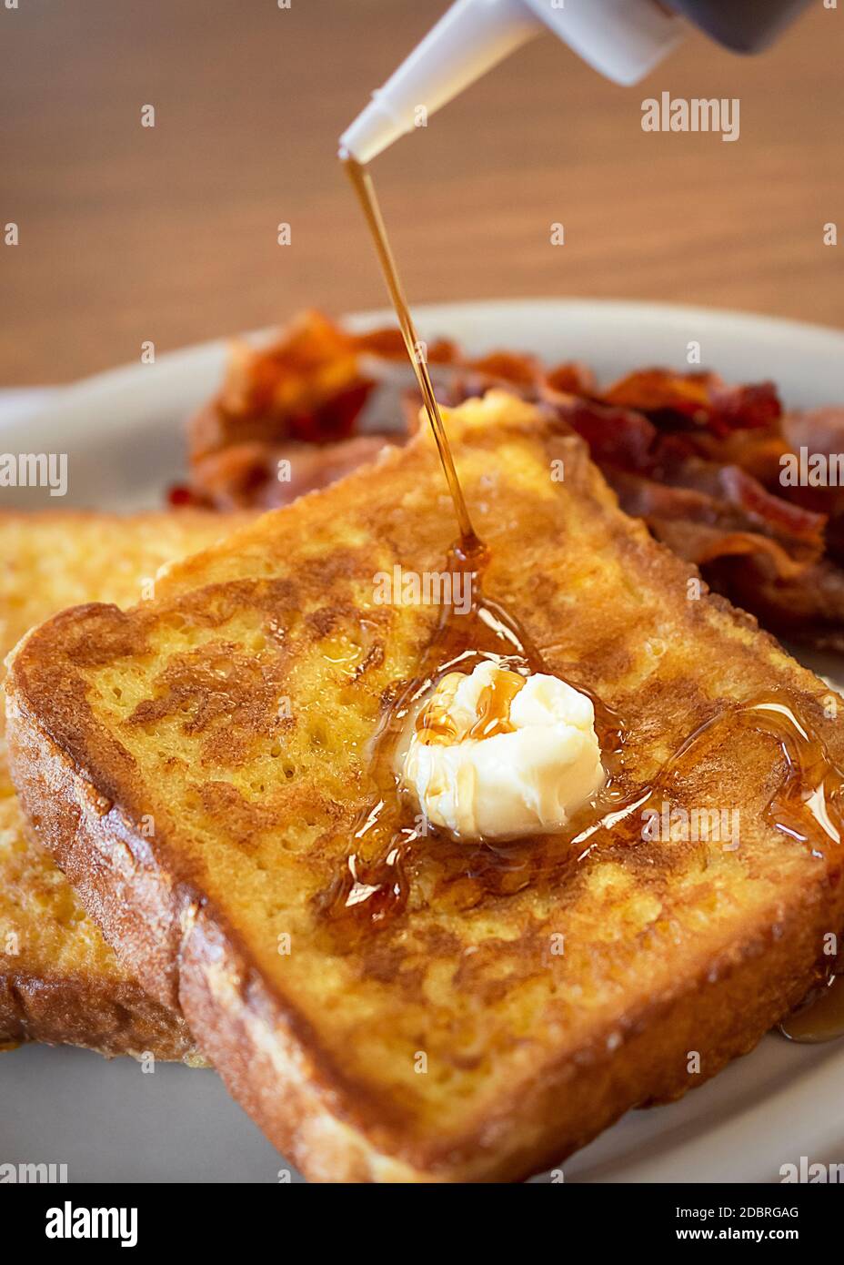 Zwei Stücke French Toast auf einem weißen Teller mit Speck verschwommen im Hintergrund. Daub Butter schmilzt auf, während Ahornsirup gegossen wird. Phot Stockfoto