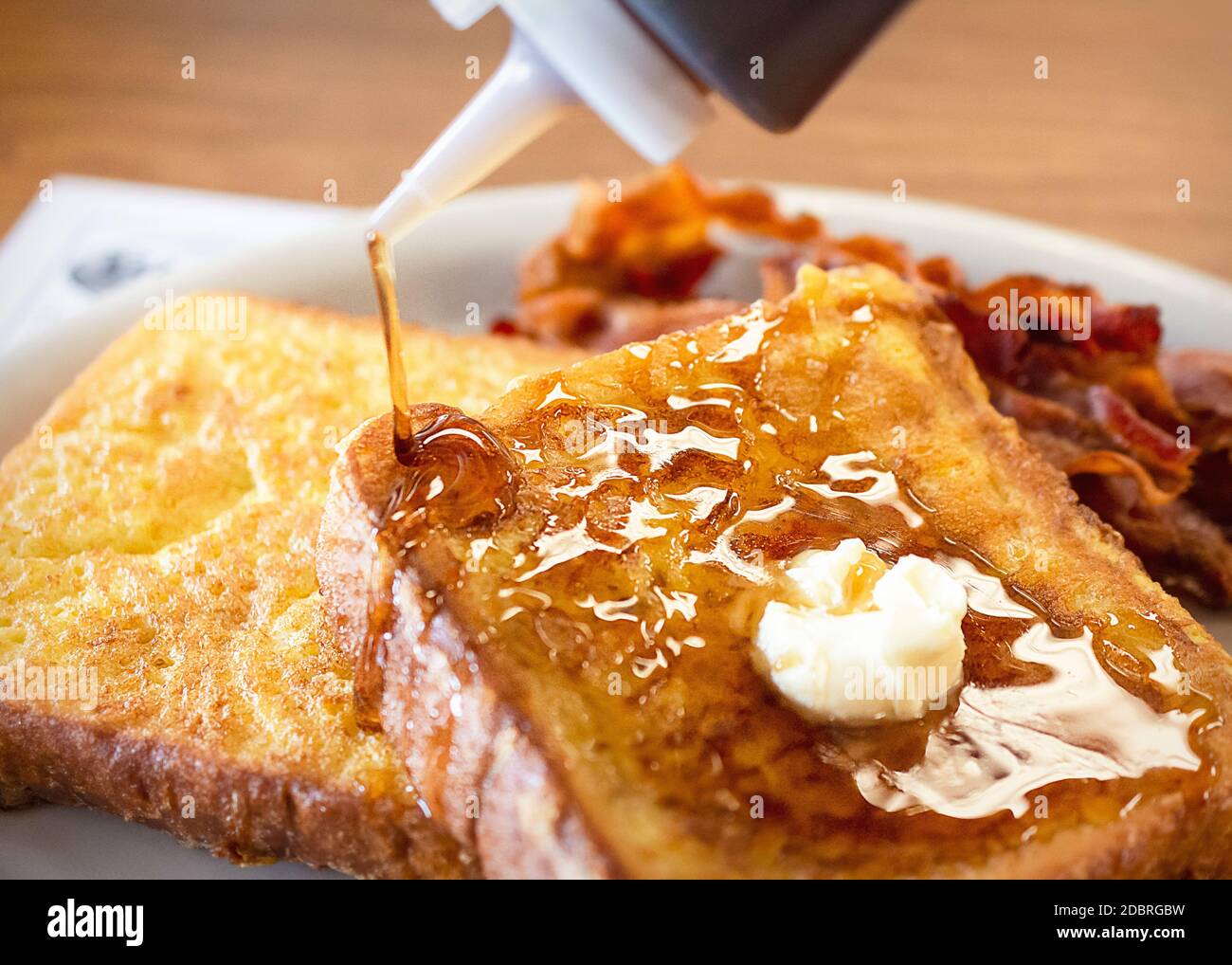 Zwei Stücke French Toast auf einem weißen Teller mit Speck verschwommen im Hintergrund. Daub Butter schmilzt auf, während Ahornsirup gegossen wird. Phot Stockfoto