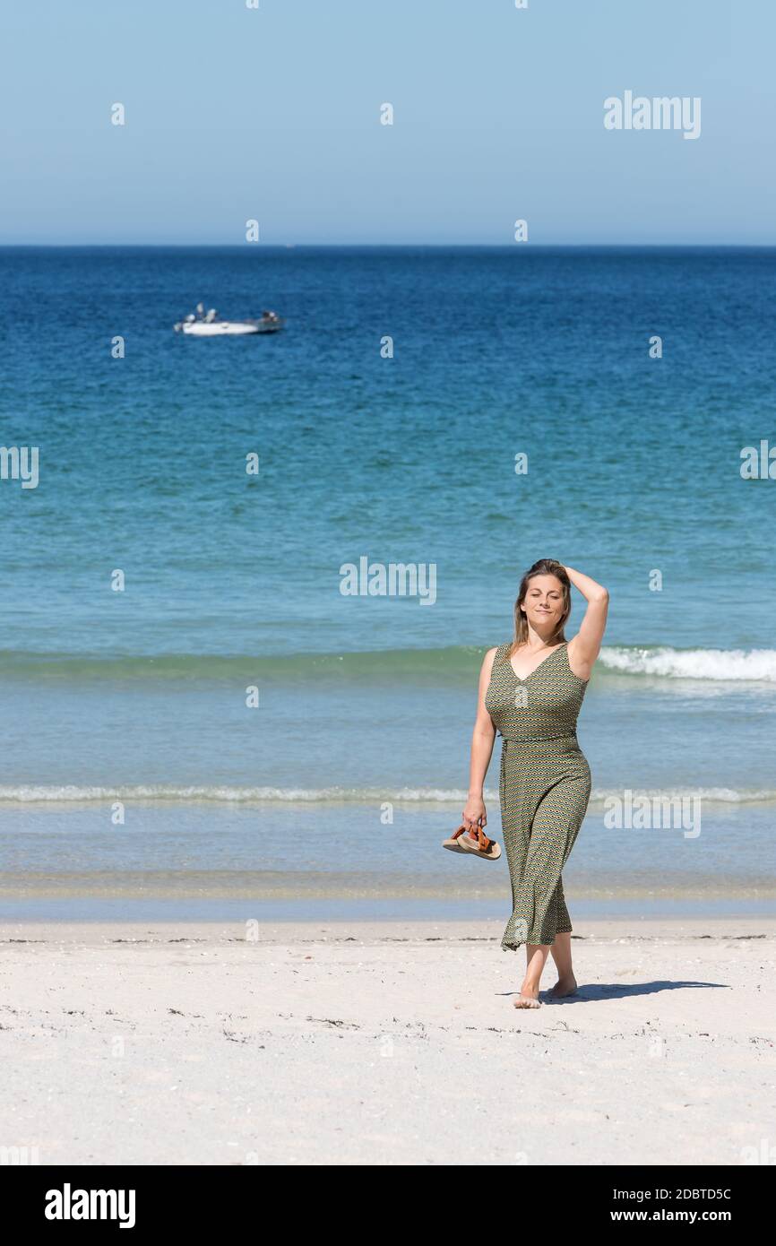 Blonde Frau mittleren Alters in einem grünen Kleid, die barfuß auf dem Weg ist Der Strand am Strand mit ihren Sandalen in einem Hand Stockfoto