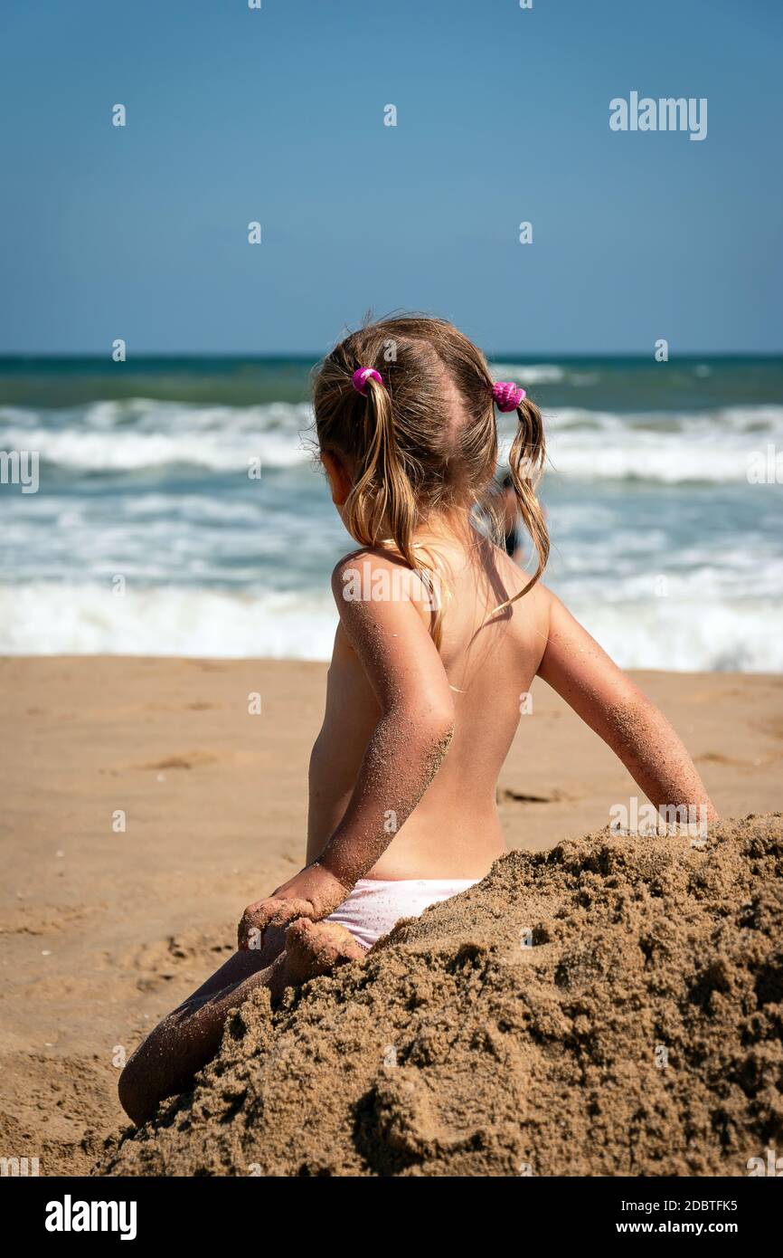 Baby Mädchen in der Sommerzeit Rückansicht. Mädchen Kind entspannen allein am Meer. Vorschulblond, die auf die Wellen des Ozeans schaut. Liebenswert kleines Kind zurück sitzen an Stockfoto