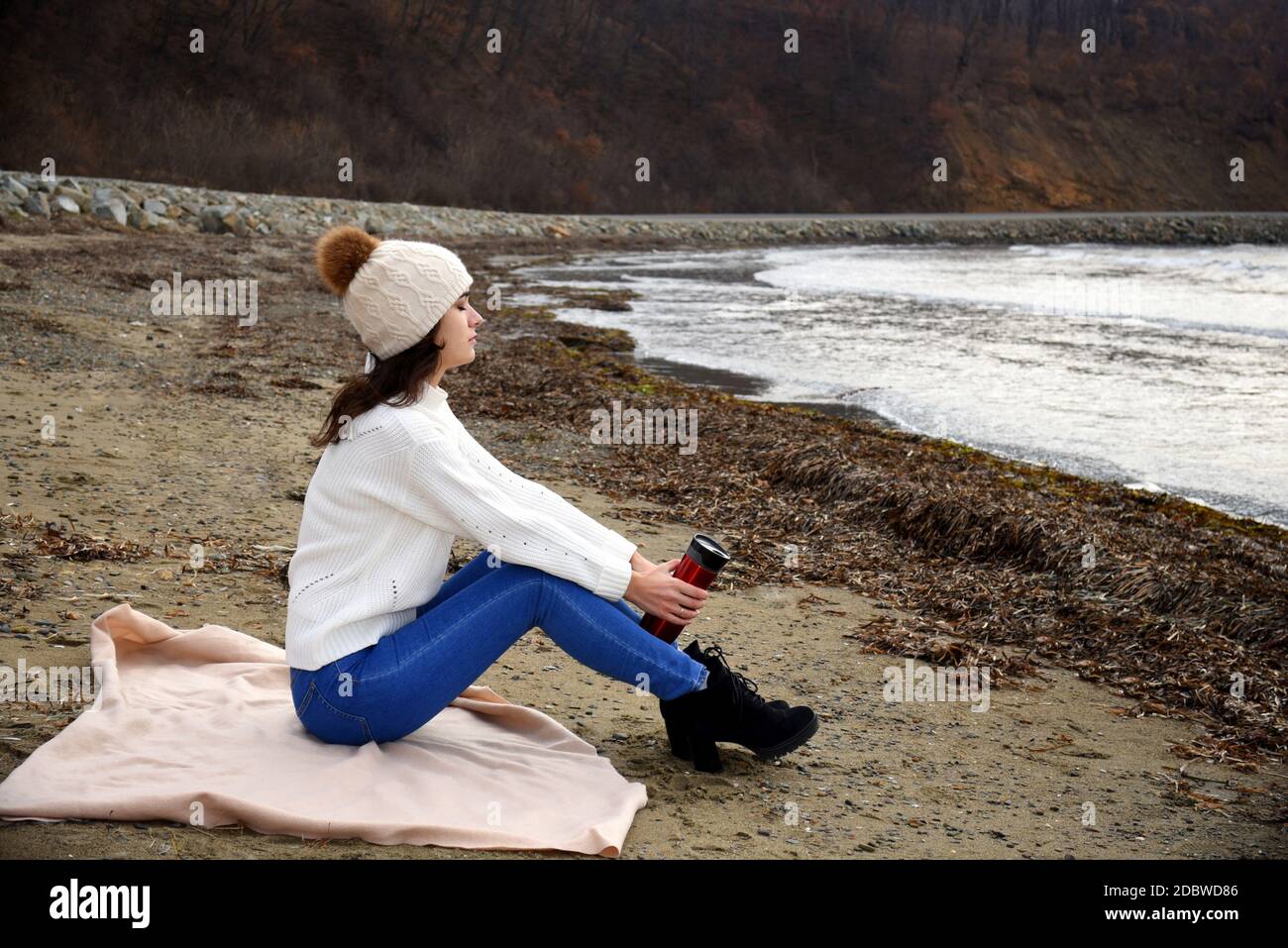 Junge Frau in weißem Pullover, Hut mit Pom-Pom, sitzt am Ufer des japanischen Meeres mit geschlossenen Augen, inhalieren den Duft der Winterluft. Stockfoto