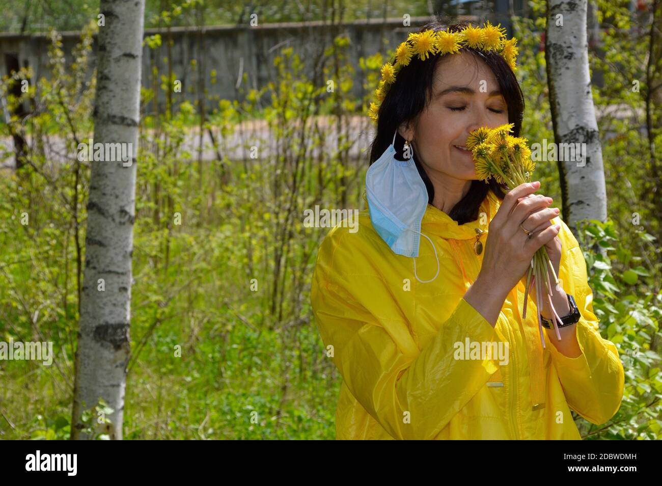 Brunette Frau mit geschlossenen Augen, in gelber Jacke mit Kranz von Löwenzahn und entfernt medizinische Maske riechenden Strauß von Löwenzahn. Coronavirus pande Stockfoto