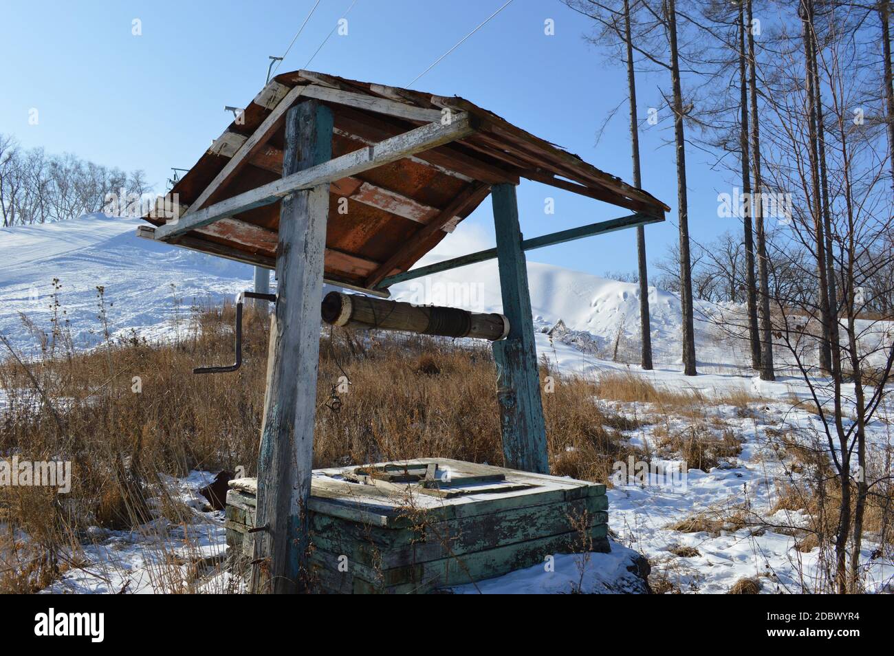 Alte verlassene hölzerne Brunnen auf dem Hintergrund des trockenen braunen Grases und des weißen Schnees des Snowboardkurortes in den Bergen, Russland. Stockfoto