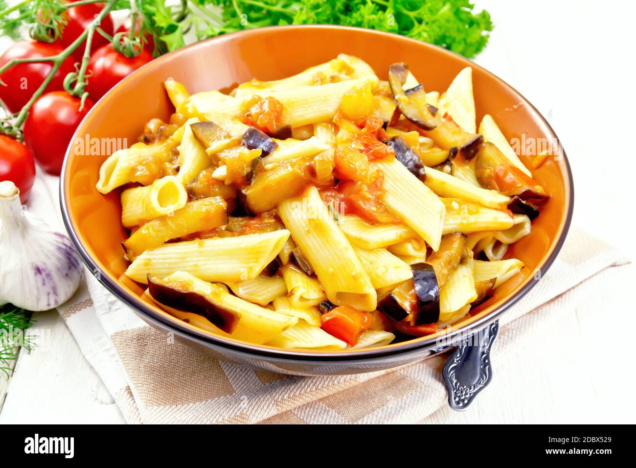 Penne Pasta mit Auberginen und Tomaten in einer Schüssel auf Küchenhandtuch, Gabel, Knoblauch und Petersilie auf hellem Holzbrett Hintergrund Stockfoto
