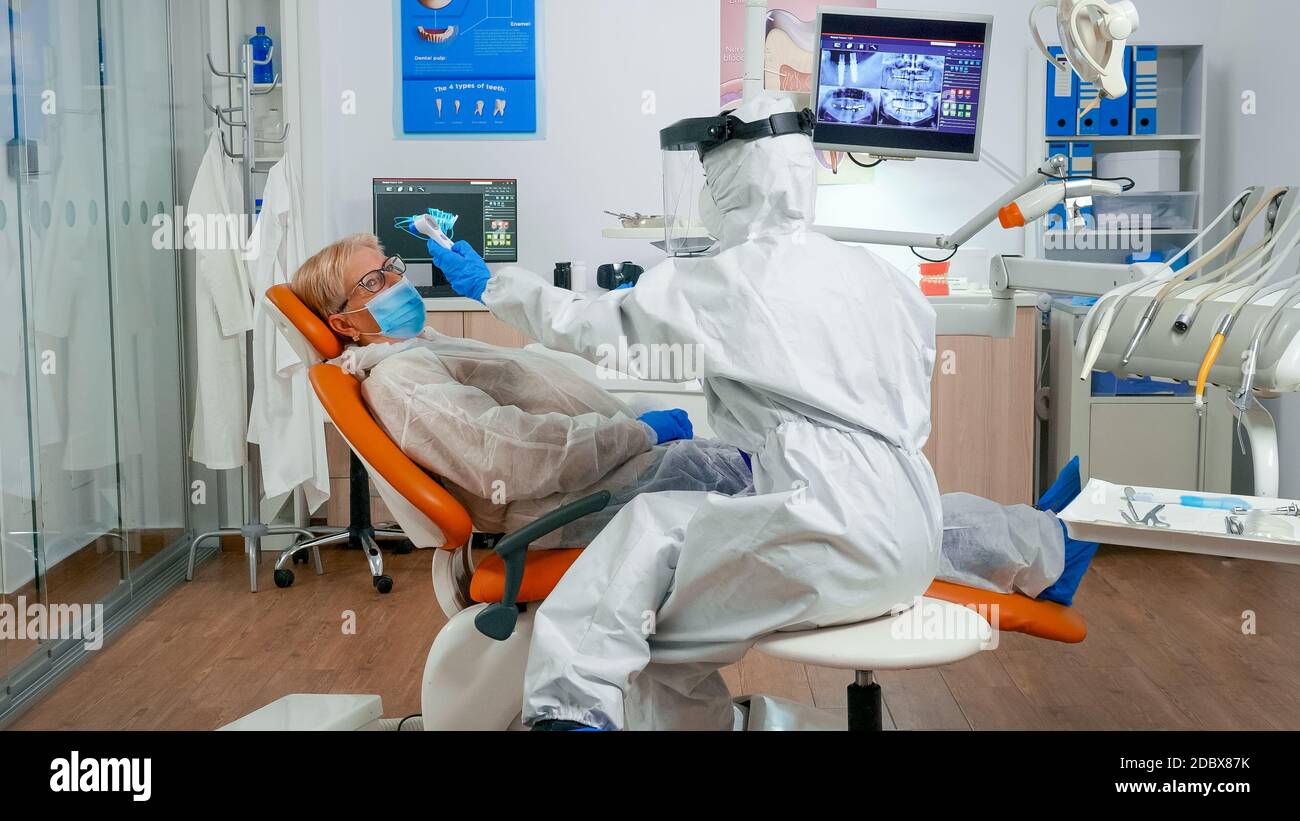Zahnarzthelfer in potective Anzug Messung der Patiententemperatur vor zahnärztliche Untersuchung während Coronavirus Pandemie. Krankenschwester im Overall und Maske verhört Frau und Notizen auf Zwischenablage Stockfoto