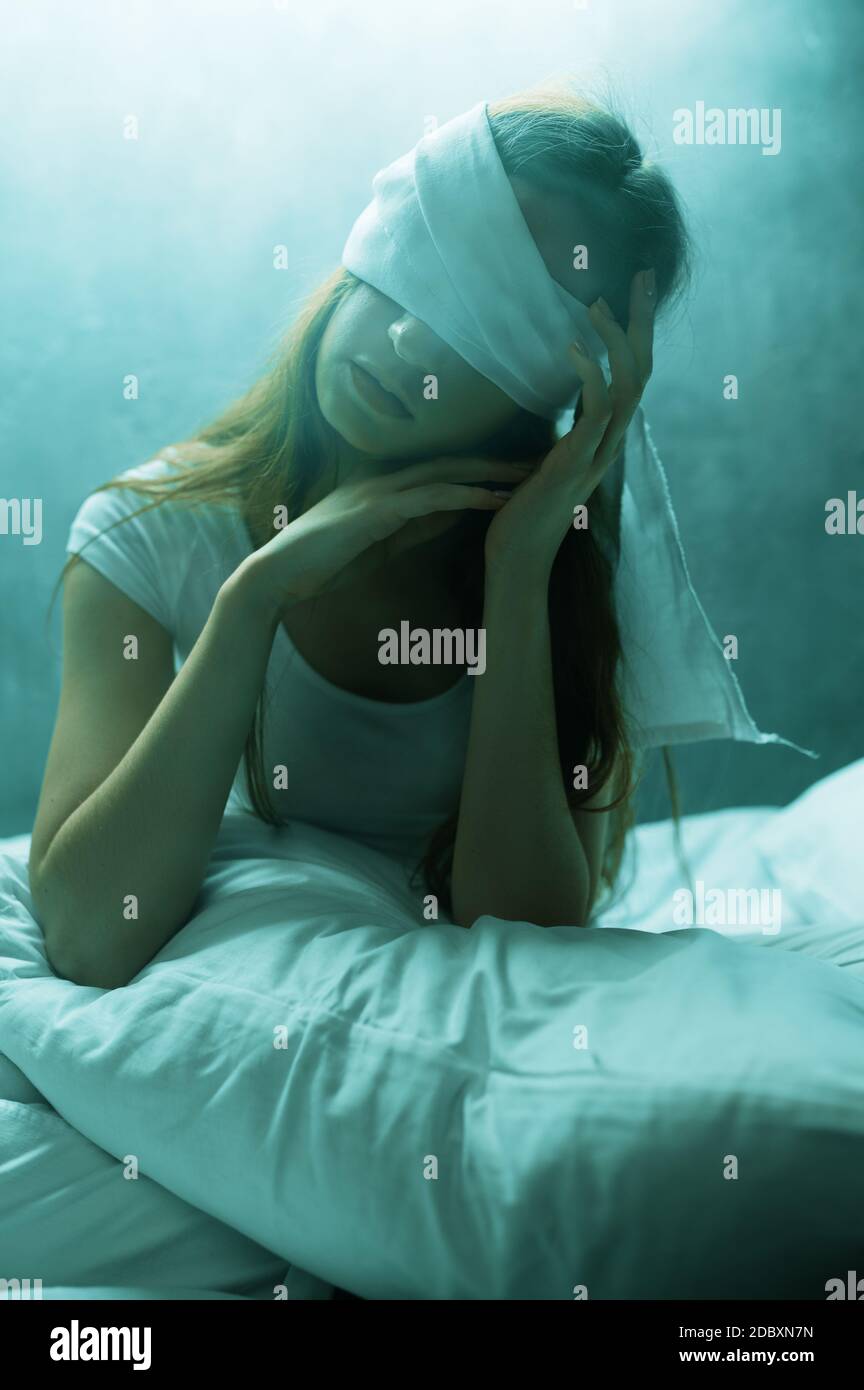 Verrückte Frau mit verbundenen Augen im Bett sitzend, dunkles Zimmer auf dem Hintergrund. Psychedelische weibliche Person mit Problemen jede Nacht, Depression und Stress, Traurigkeit Stockfoto