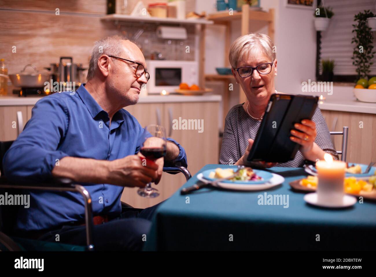 Ältere Frau, die auf einem Tablet-pc browst, und behinderter Mann im Rollstuhl, der Weinglas hält. Imobilisierter behinderter älterer Ehemann, der am Telefon surft und den festlichen Mann genießt und ein Glas Rotwein trinkt. Stockfoto