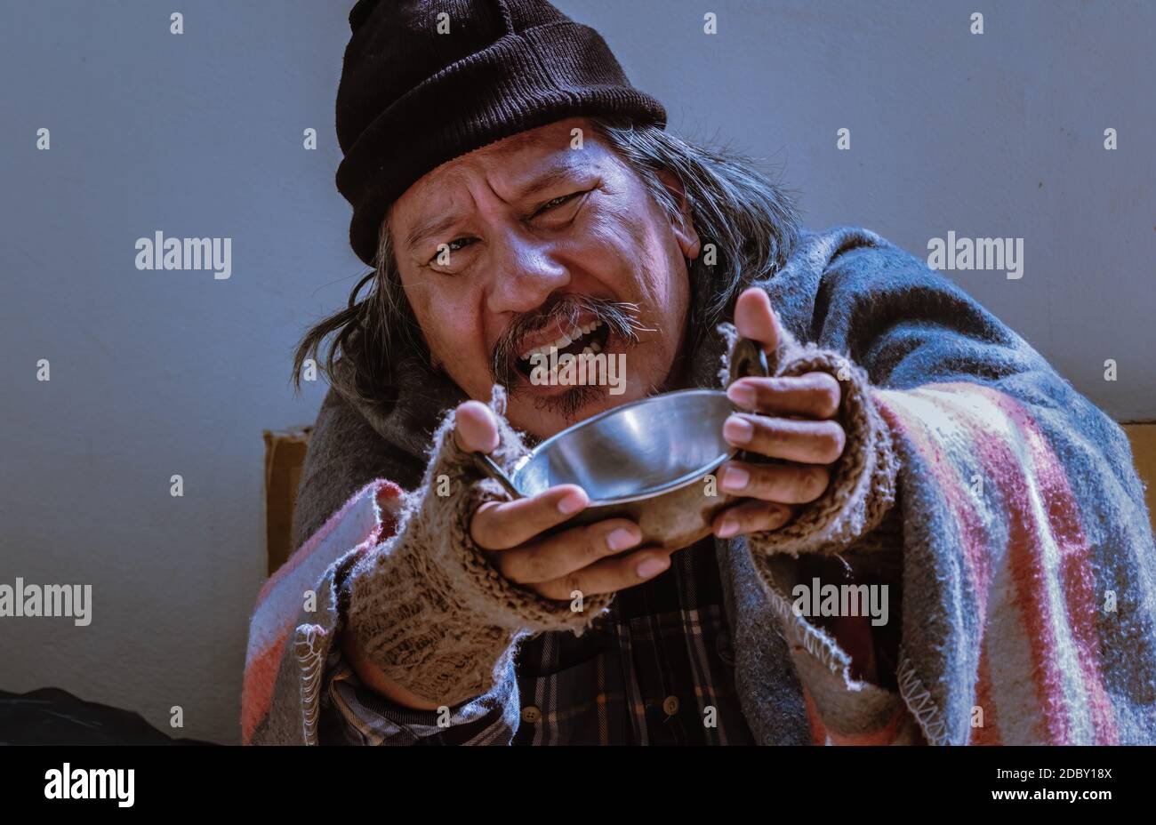 Ein erbärmlicher Obdachloser mit unglücklichem Blick bittet um Hilfe, indem er mit einer Schüssel bettelt. Stockfoto