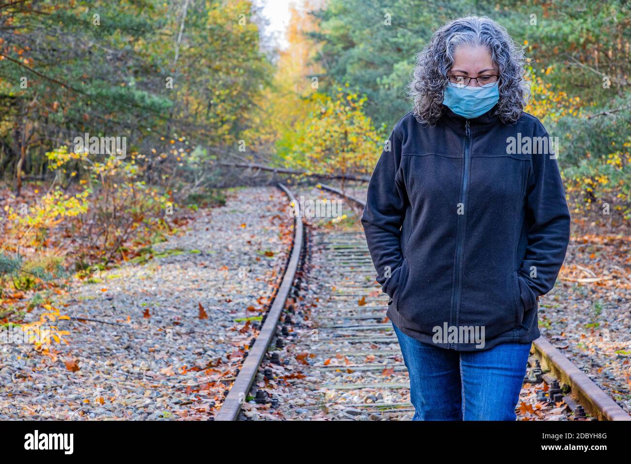 Reife Frau, grauschwarzes Haar mit Maske, sehr nachdenklich auf stillem Bahngleis, alte Eisenrheinbahn (IJzeren Rijn), Herbsttag in Meinweg n Stockfoto