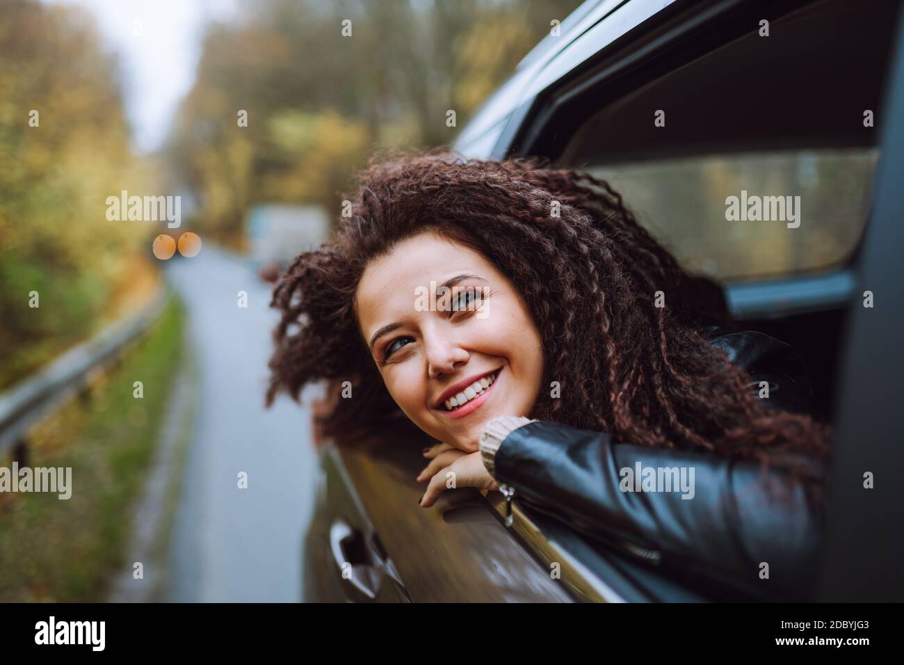 Junge afro Haar Frau reisen mit dem Auto auf wilden Wald Herbst Straße. Weiblicher Blick im offenen Fenster von hinten sitzen mit glücklichem Lächeln. Stockfoto