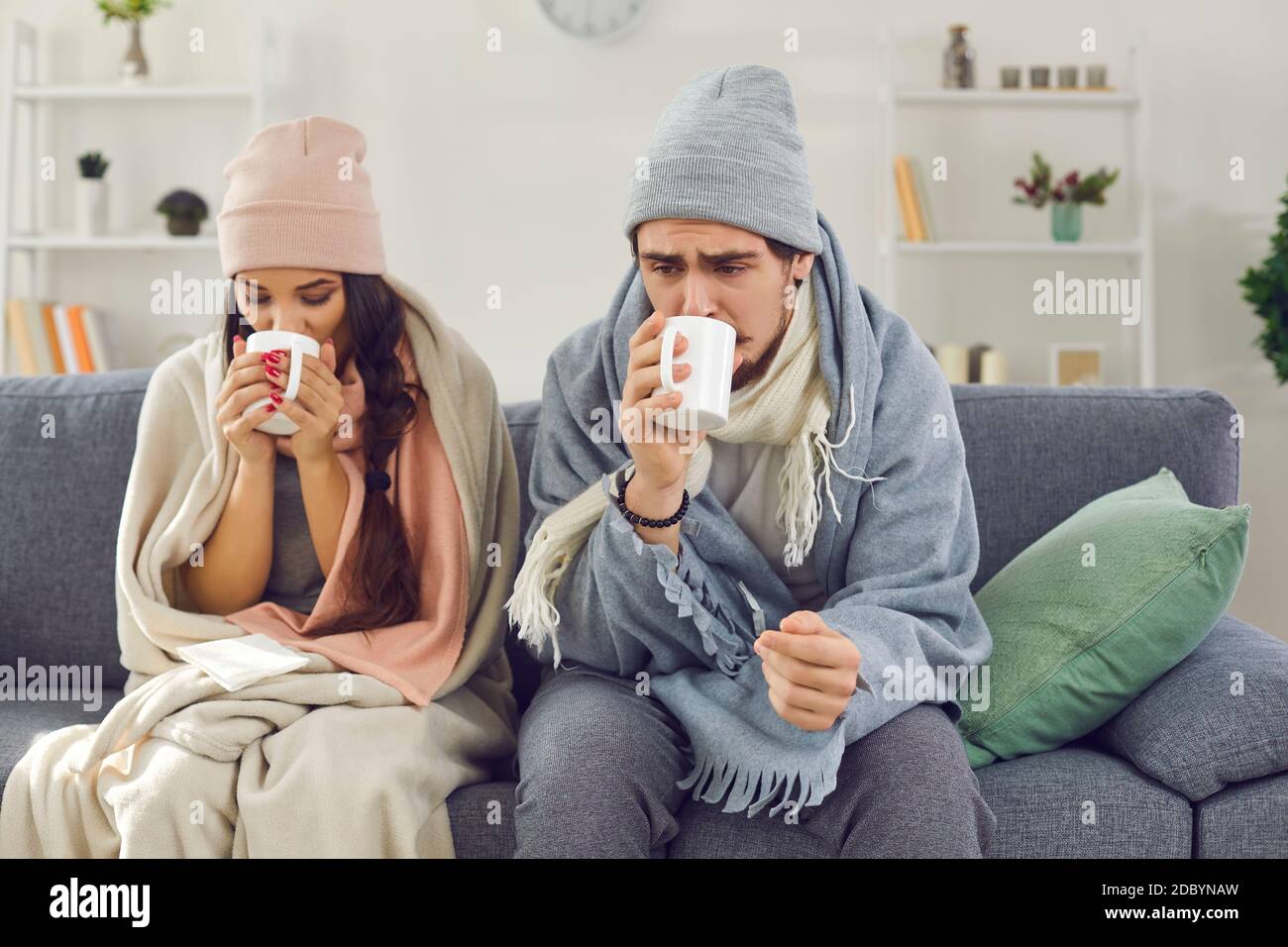 Kranke junge Paar in Plaids eingewickelt sitzen auf dem Sofa, trinken heißen Tee und versuchen, sich aufzuwärmen Stockfoto