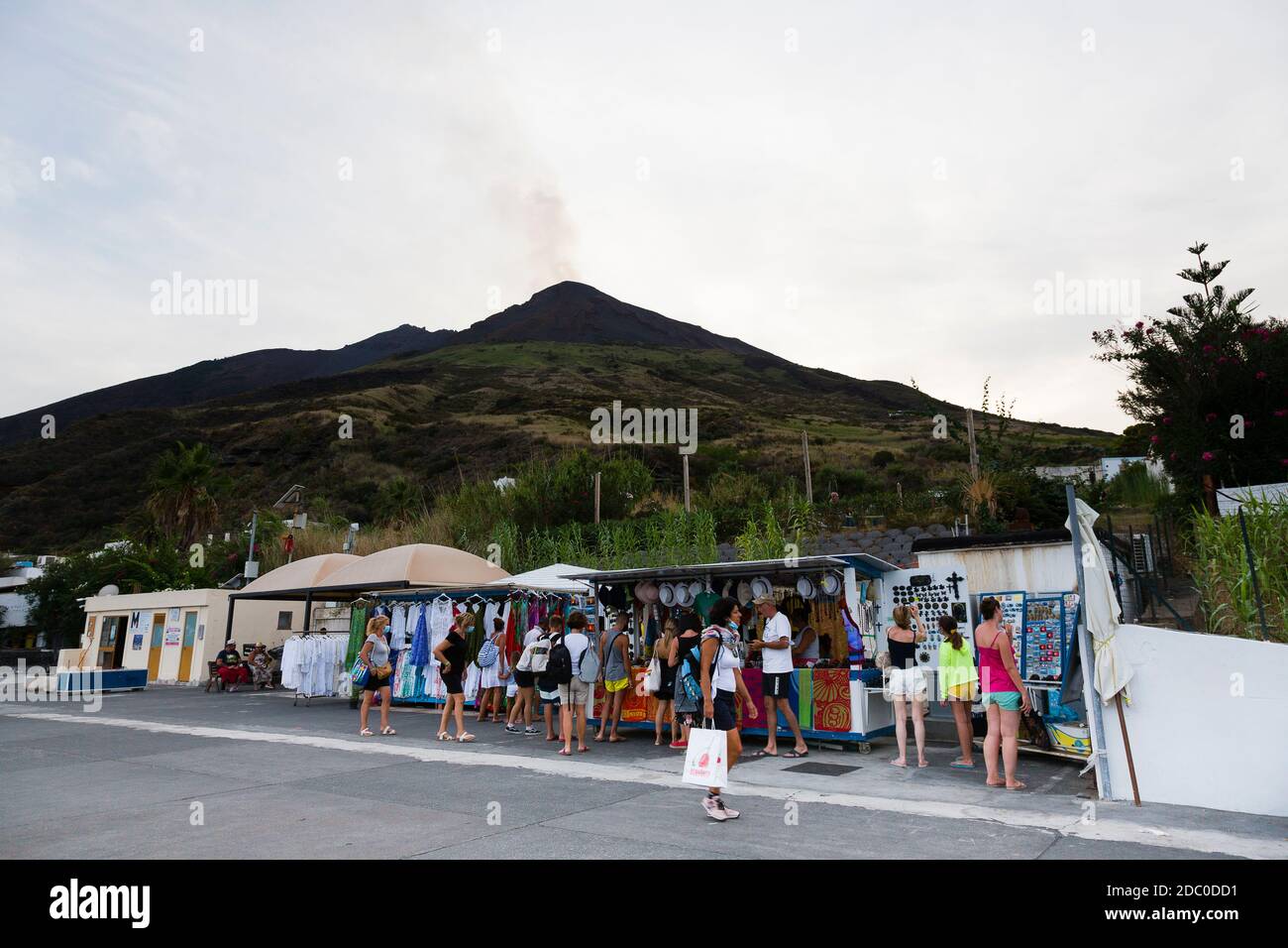 Sizilien, Italien. Touristen wandern entlang einer der Hauptstraßen der vulkanischen Insel Stromboli, vorbei an kleinen Hütten, die Souvenirs verkaufen. Der Vulkan von Stro Stockfoto