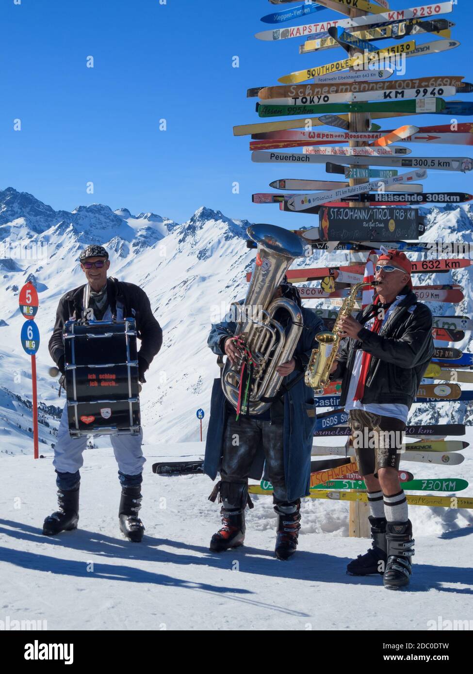 Musikkapelle in Karnevalskostümen spielt im Skigebiet Ischgl-Samnaun mit Skistäste, die auf internationale Städte dahinter, Österreich, verweisen Stockfoto