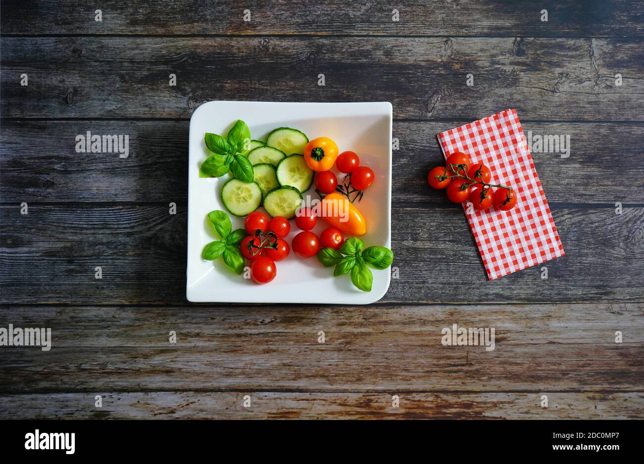 Frische Zutaten für einen gesunden Salat auf einem weißen quadratischen Teller: Tomaten, Paprika, Zucchini und Basilikum. Dunkler Holzhintergrund mit karierter Serviette. Stockfoto