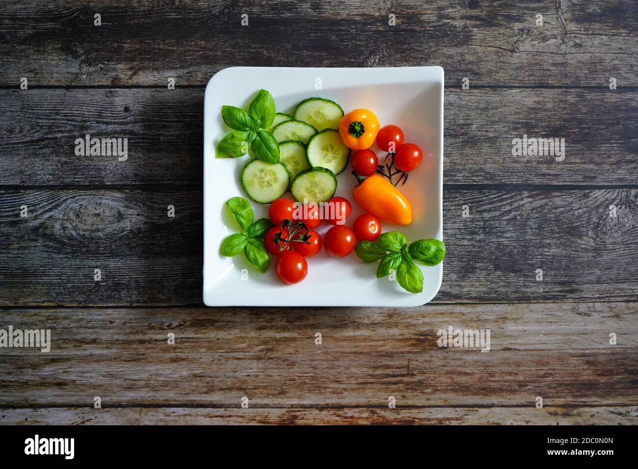 Frische Zutaten für einen gesunden Salat auf einem weißen quadratischen Teller: Tomaten, Paprika, Zucchini und Basilikum. Dunkler Holzhintergrund. Stockfoto