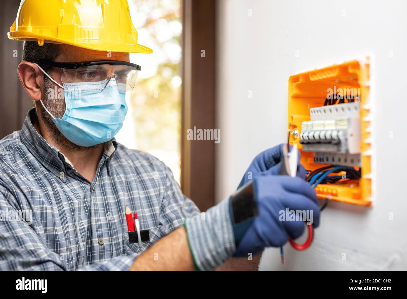 Elektriker bei der Arbeit an einem elektrischen Panel mit Helm, Schutzbrille und Handschuhe geschützt; tragen Sie die chirurgische Maske, um die Ausbreitung von Coronavirus zu verhindern Stockfoto