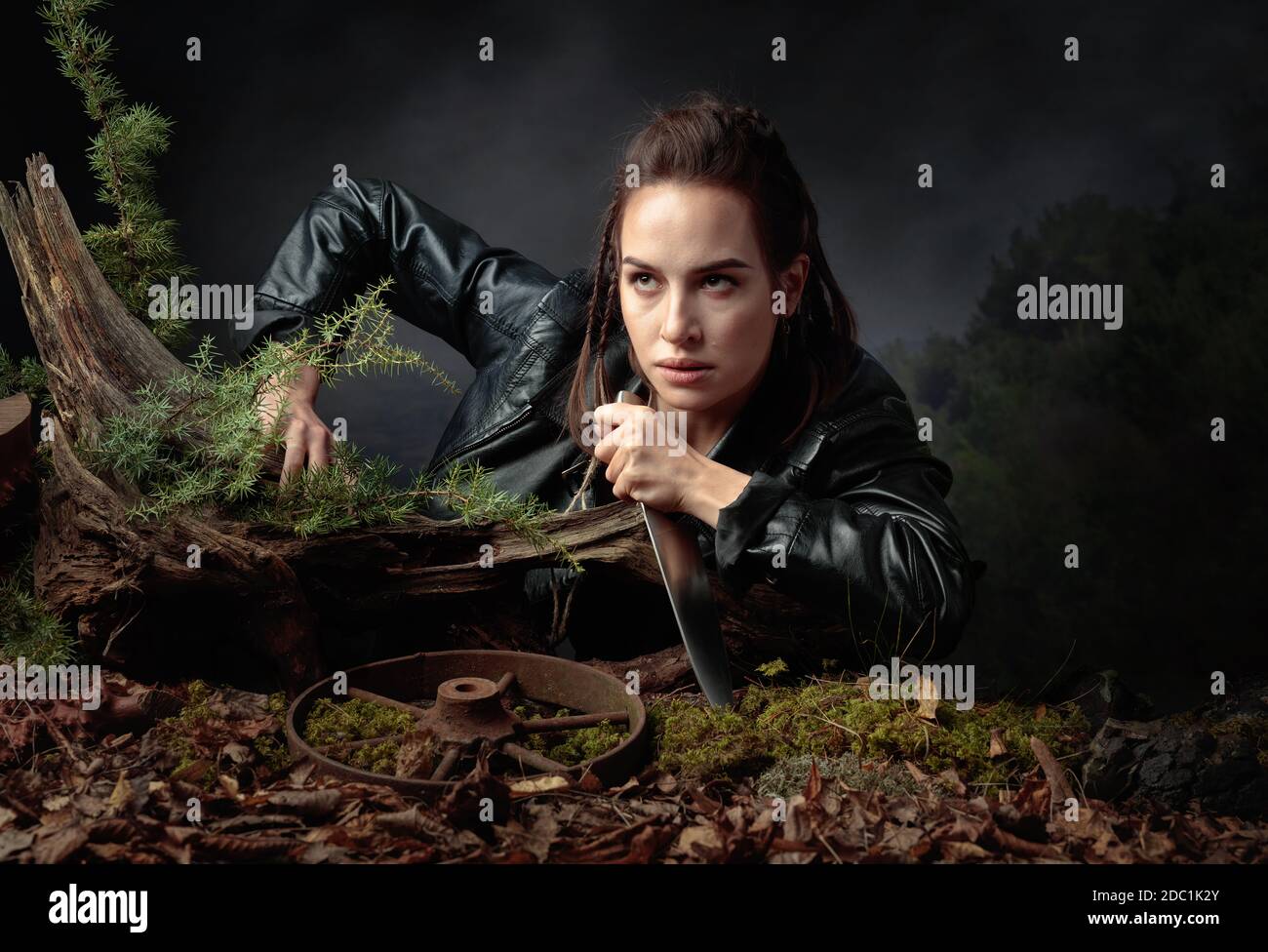 Porträt einer jungen Frau mit einem Messer. Ein seriöses Mädchen in einer schwarzen Lederjacke mit einem Messer klettert aus der Grube im Wald. Stockfoto