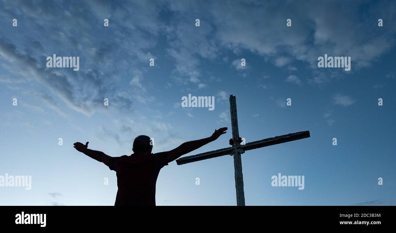 Mann mit ausgestreckten Armen, der unter dem Kreuz auf dem Gipfel des Berges betet. Stockfoto
