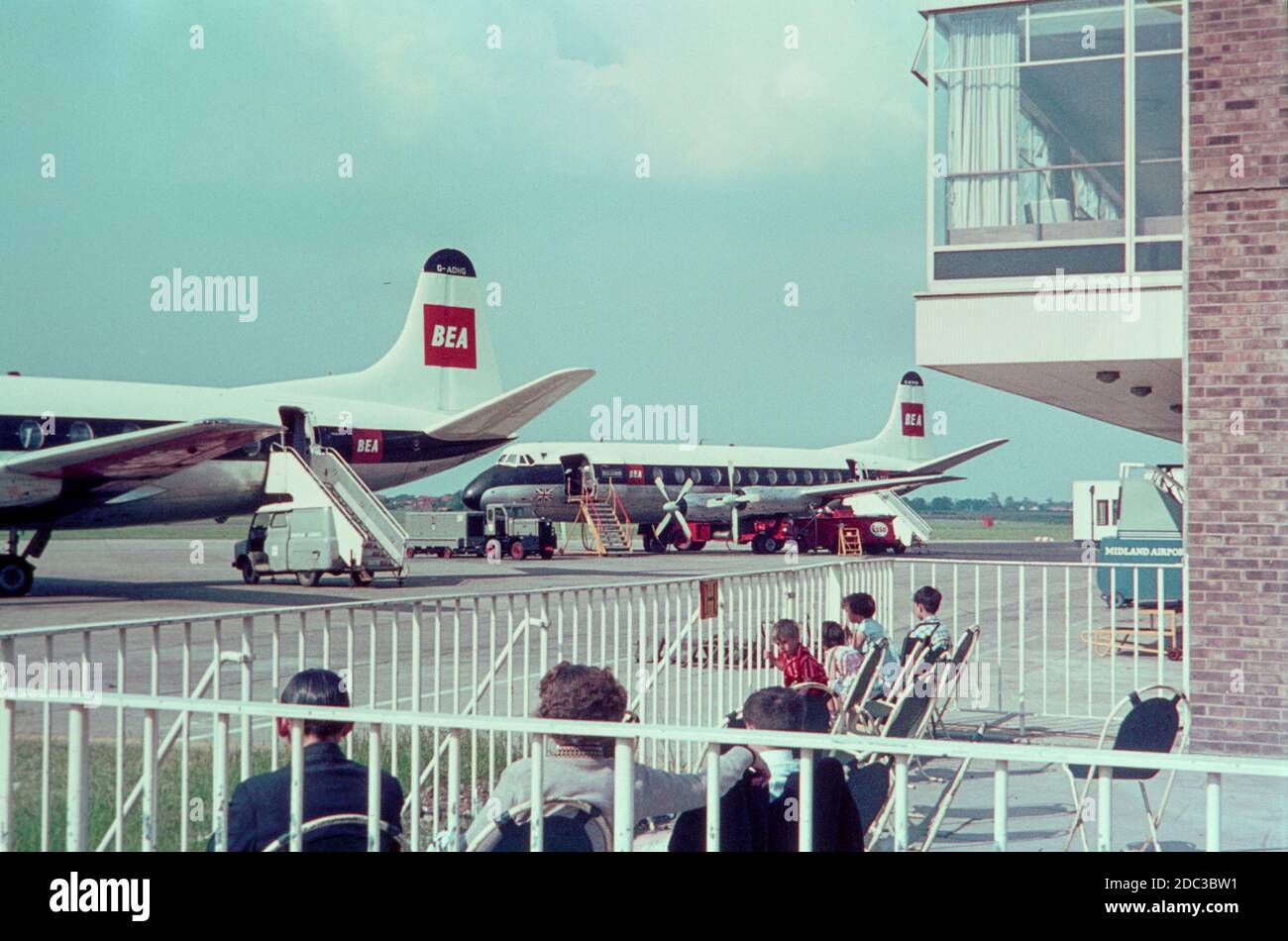 Ein Vintage-Farbfoto, aufgenommen in den 1960er Jahren am Birmingham Airport in England. Zeigt zwei Flugzeuge der British European Airways, BEA, Vickers Viscount, einschließlich G-AOHG, vor dem Terminalgebäude, die auf ihre Passagiere warten. Im Vordergrund sitzt eine Gruppe von Menschen, die das Flugzeug beobachten. Stockfoto