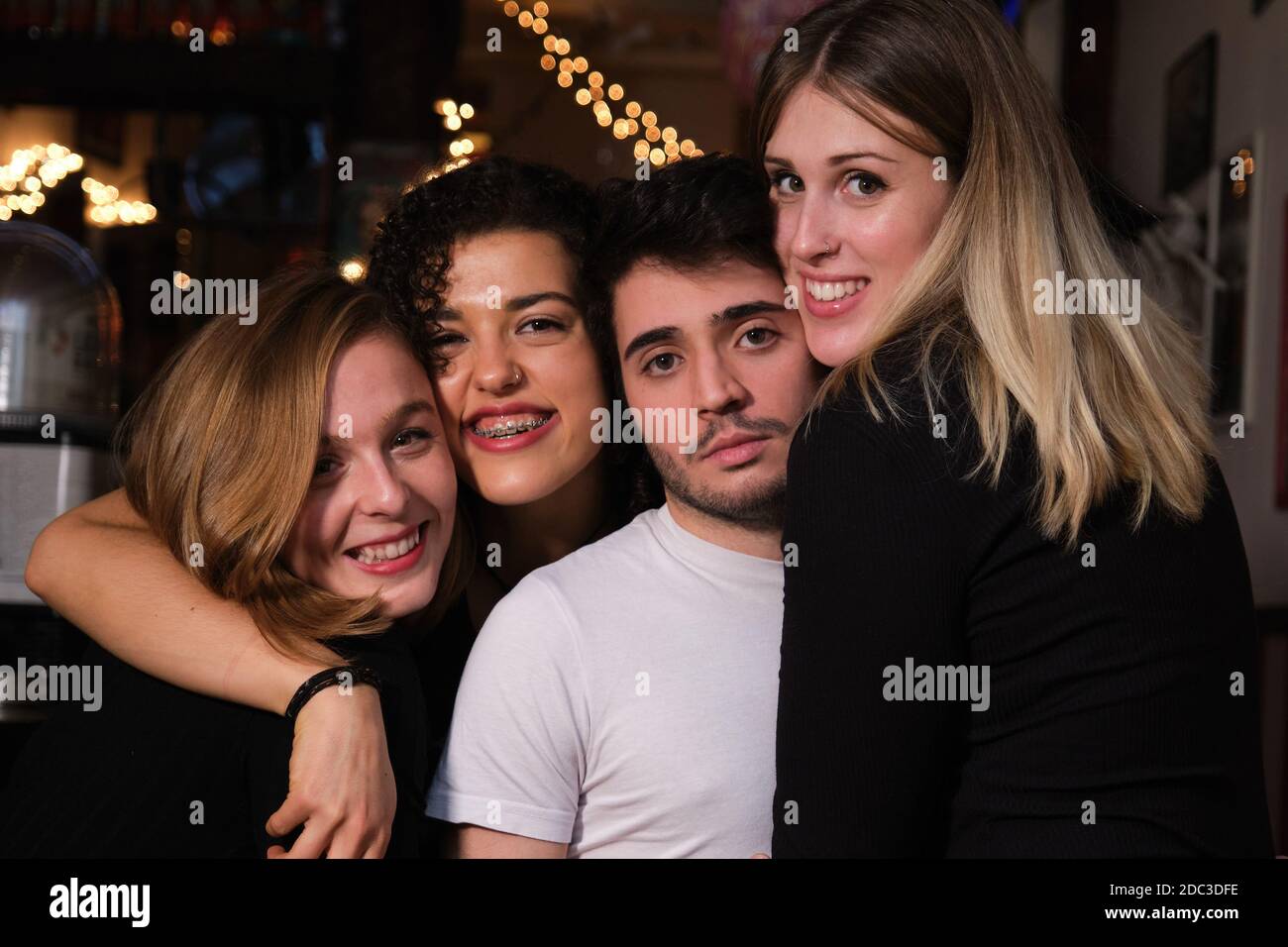 Drei lächelnde junge Frauen und ein ernsthafter junger Mann umarmten sich gegenseitig in einer Bar. Gruppe von Freunden. Stockfoto