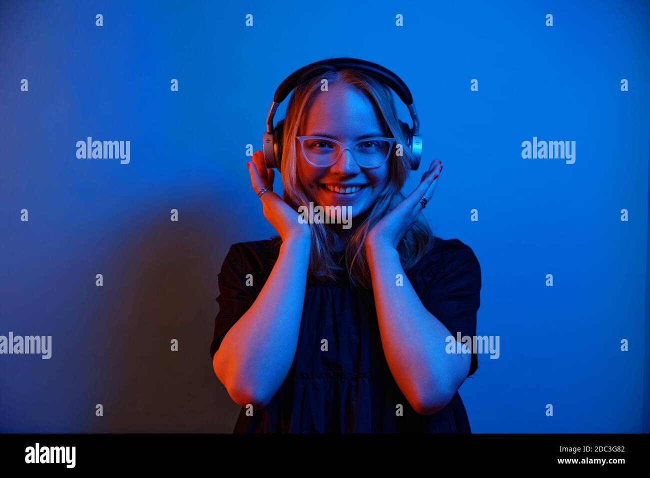 Junge kaukasische Mädchen trägt Brille und Kopfhörer schaut auf die Kamera und lächelt auf einem blauen Neon Hintergrund. Stockfoto