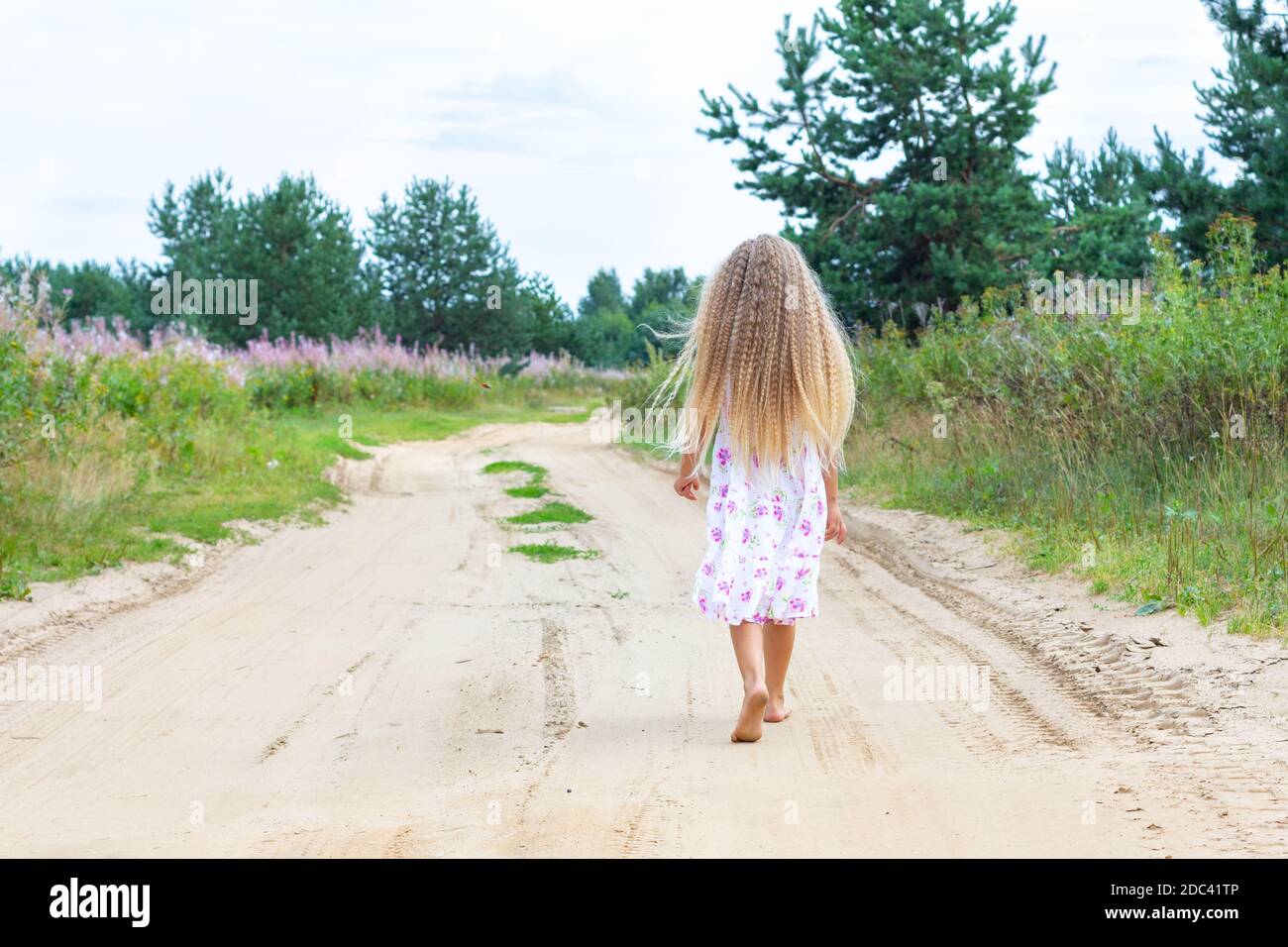 Ein Mädchen mit langen blonden lockigen Haaren geht entlang einer ländlichen Straße. Porträt von hinten in voller Länge. Ein Spaziergang an der frischen Luft in Einsamkeit. Stockfoto