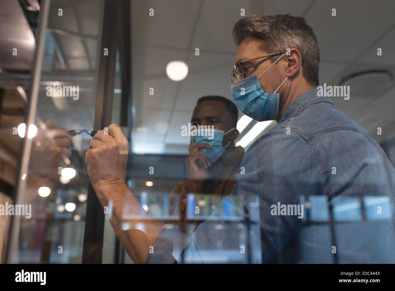 Kaukasischer Mann trägt Gesichtsmaske schreiben mit Marker Stift auf Glasplatine im modernen Büro Stockfoto