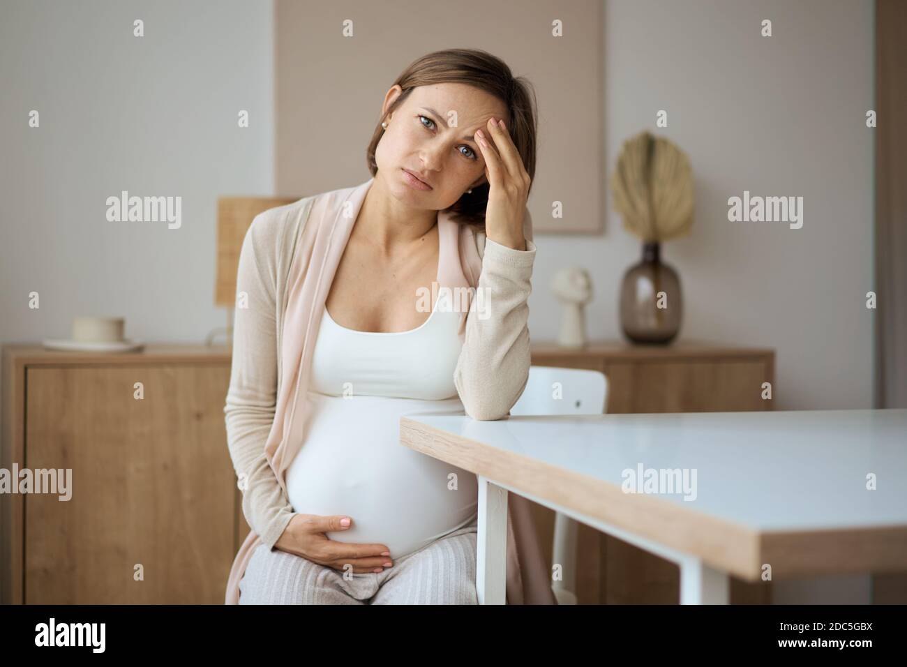 Schwangere Frau, die an Kopfschmerzen leidet, während sie am Tisch sitzt Stockfoto