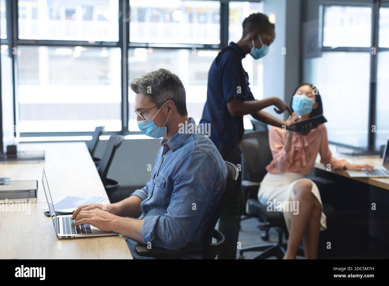 Kaukasischen Mann trägt Gesichtsmaske mit Laptop, während african american Frau und asiatische Frau tragen fa Stockfoto