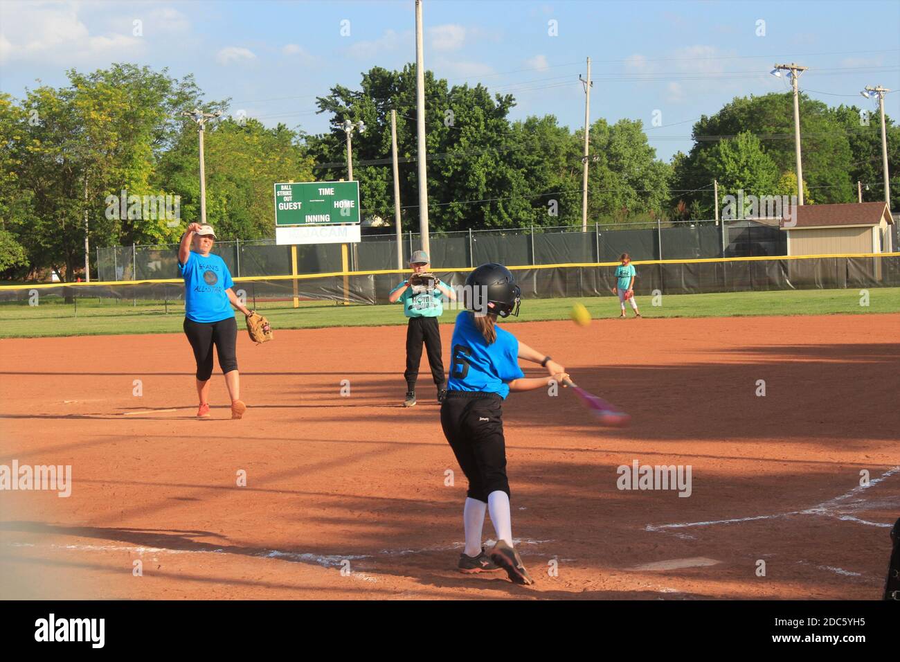 Mädchen spielen Softball mit blauem Himmel und Bäume mit rotem Schmutz auf dem Spielfeld in Kansas. Stockfoto