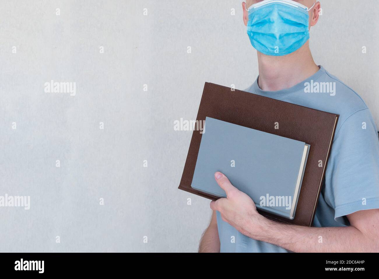 Ein junger Mann in einer OP-Maske und einem blauen T-Shirt hält ein Hardcover und ein Lederbuch auf einem texturierten Stoffhintergrund. Stockfoto