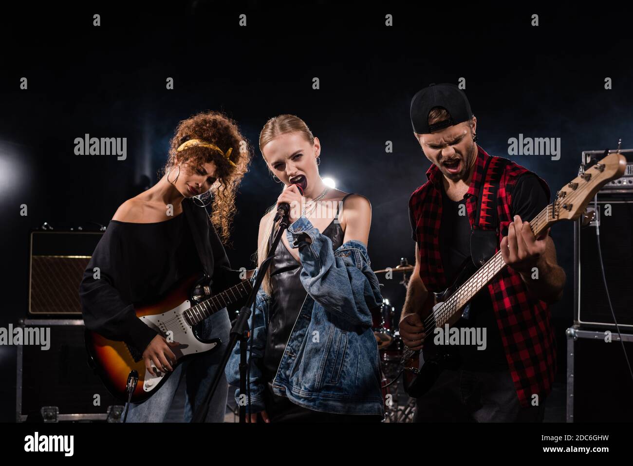 KIEW, UKRAINE - 25. AUGUST 2020: Blonde Sängerin der Rockband singt neben Musikern mit Bass-Gitarren mit Hintergrundbeleuchtung auf schwarz Stockfoto