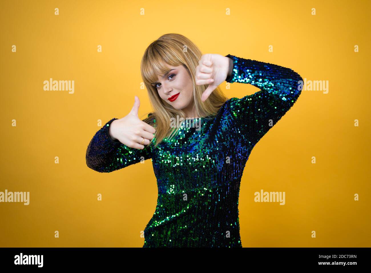 Schöne blonde Frau trägt ein ausgefallenes Kleid über gelben Hintergrund tut Daumen nach oben und unten, Meinungsverschiedenheiten und Übereinkunft Ausdruck. Verrückter Konflikt Stockfoto